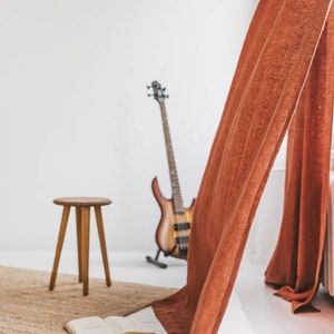 May include: A brown bass guitar leans against a white wall, with a brown stool and a book on a white wooden floor. A rust-colored curtain hangs in the background.