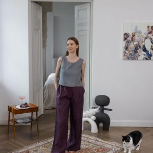 K&ouml;nnte beinhalten: Eine Frau tr&auml;gt ein graues &auml;rmelloses Oberteil und eine dunkelviolette Hose mit weitem Bein. Das Outfit wird mit einem gemusterten Teppich und einer schwarz-wei&szlig;en Katze kombiniert. Der Raum hat ein minimalistisches Design mit wei&szlig;en W&auml;nden und Holzb&ouml;den.