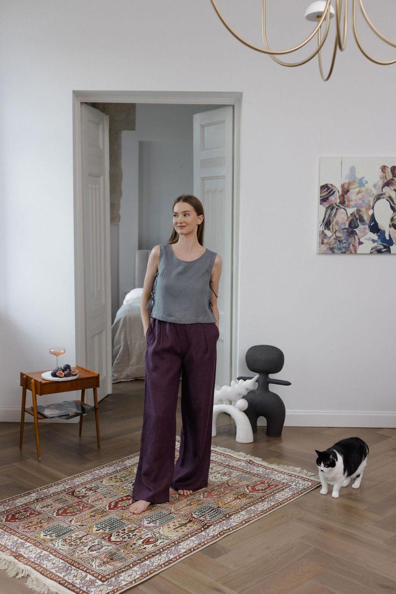 K&ouml;nnte beinhalten: Eine Frau tr&auml;gt ein graues &auml;rmelloses Oberteil und eine dunkelviolette Hose mit weitem Bein. Das Outfit wird mit einem gemusterten Teppich und einer schwarz-wei&szlig;en Katze kombiniert. Der Raum hat ein minimalistisches Design mit wei&szlig;en W&auml;nden und Holzb&ouml;den.