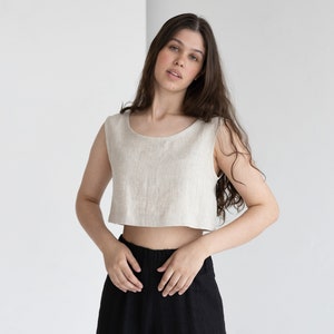 May include: A sleeveless, cropped top in a natural linen color is paired with black, wide-leg pants. The top has a scoop neckline and a relaxed fit. The pants have an elastic waistband. The outfit is simple and minimalist.