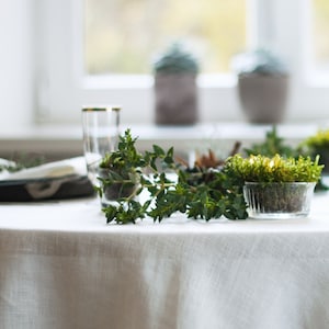 May include: A white tablecloth with a sprig of green foliage and a small glass bowl filled with moss and small lights.