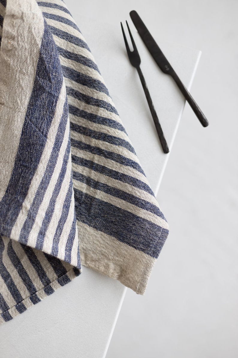 May include: A blue and white striped linen kitchen towel folded on a white surface with two black metal utensils, a fork and a knife.