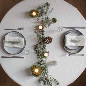 May include: A white tablecloth with two place settings, each with a gray plate, a white napkin, and silverware. A long garland of greenery with small candles in moss nests runs down the center of the table.