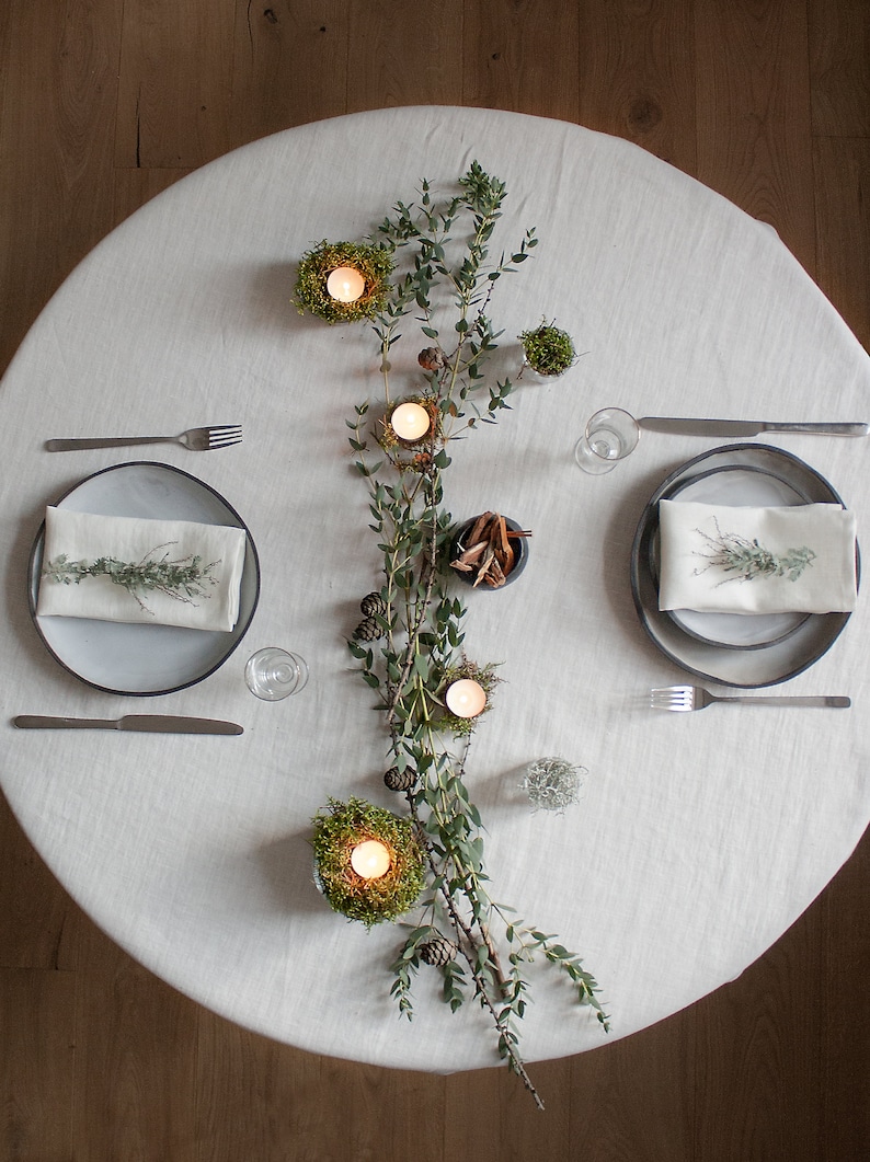 May include: A white tablecloth with two place settings, each with a gray plate, a white napkin, and silverware. A long garland of greenery with small candles in moss nests runs down the center of the table.