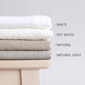 Pode incluir: Uma pilha de tecidos de linho dobrados em branco, branco sujo, natural e natural claro. Os tecidos estão colocados sobre uma superfície de madeira clara. Etiquetas de texto indicam cada cor.