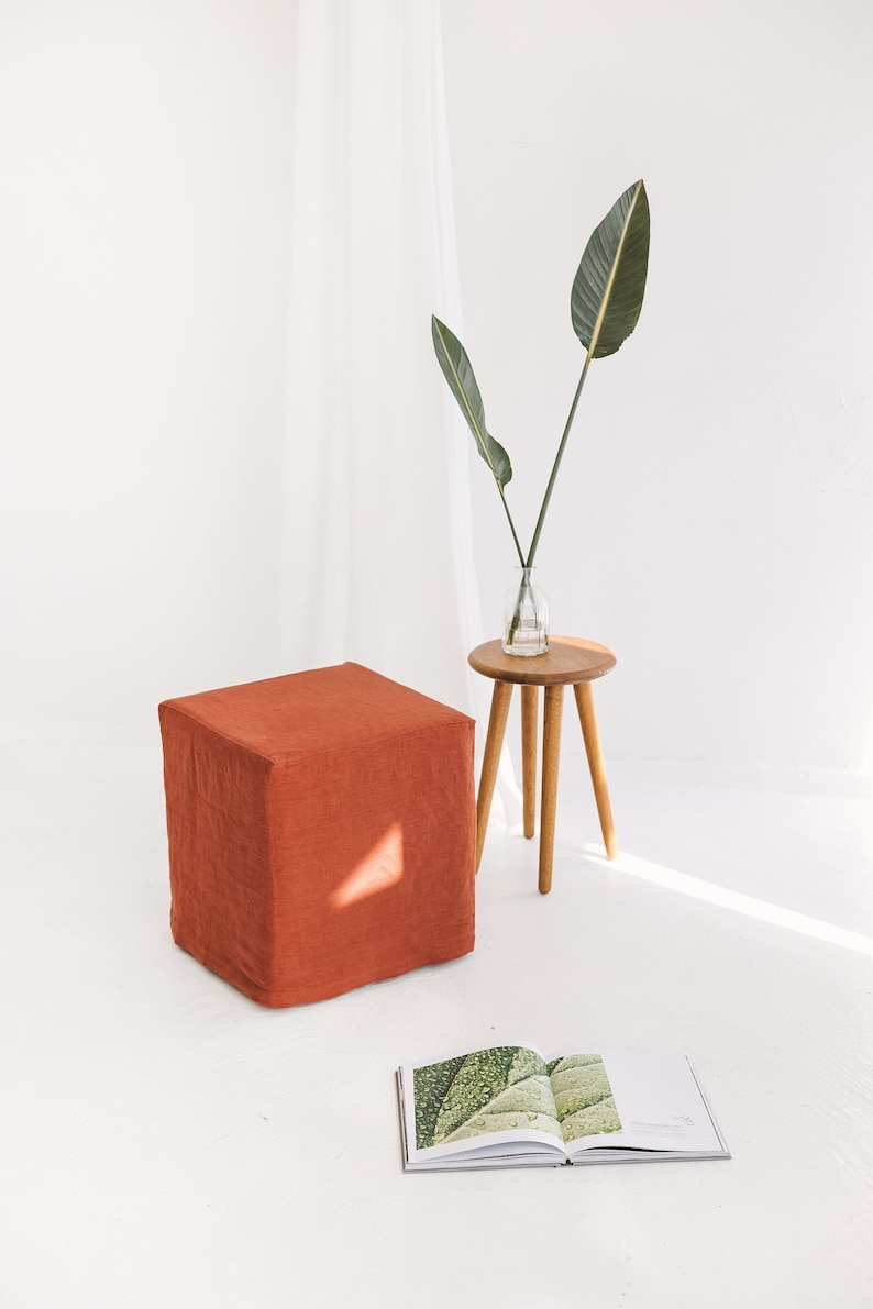 May include: A burnt orange square ottoman with a linen cover sits on a white floor. A small wooden side table with three legs holds a vase with green leaves. An open book with a green leaf image lies on the floor.