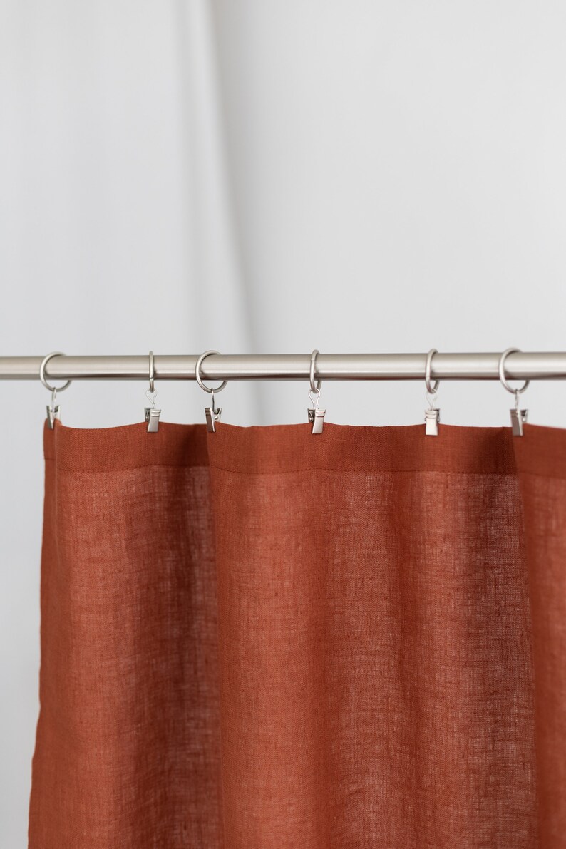 May include: A rust-colored linen curtain hangs from a silver curtain rod with silver clips.