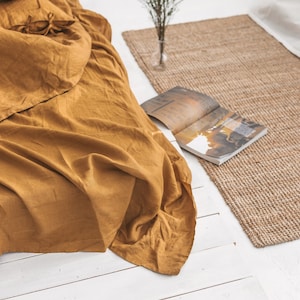 Peut inclure: Une housse de couette en lin jaune moutarde avec des liens aux coins, drap&eacute; sur un lit plateforme en bois blanc. Un petit bouquet de fleurs blanches dans un vase en verre se trouve sur un tapis en fibres naturelles.