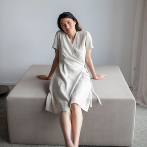 May include: A woman wearing a white linen wrap dress sits on a light gray cushioned bench. She is wearing white sandals and has her legs crossed.