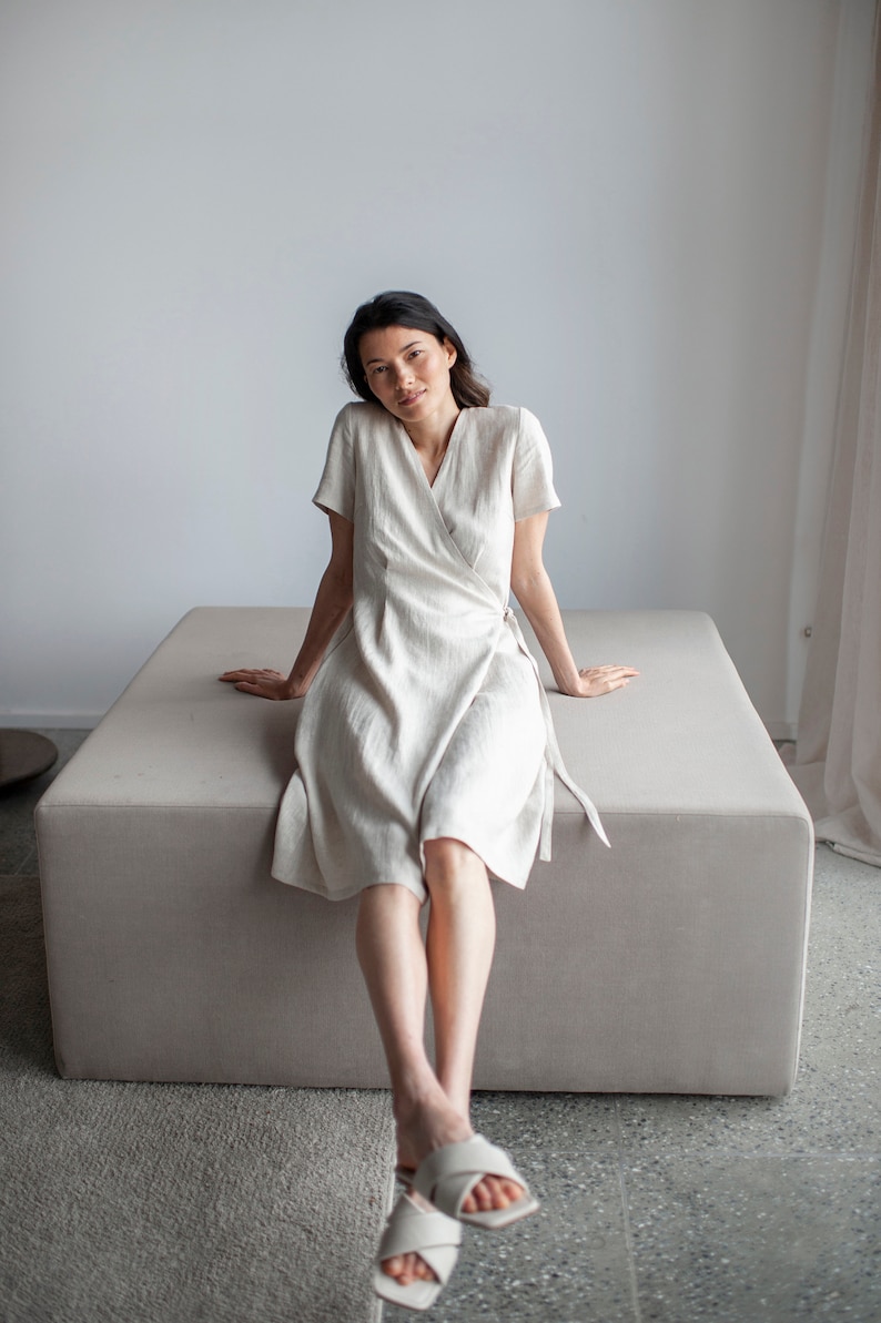 May include: A woman wearing a white linen wrap dress sits on a light gray cushioned bench. She is wearing white sandals and has her legs crossed.