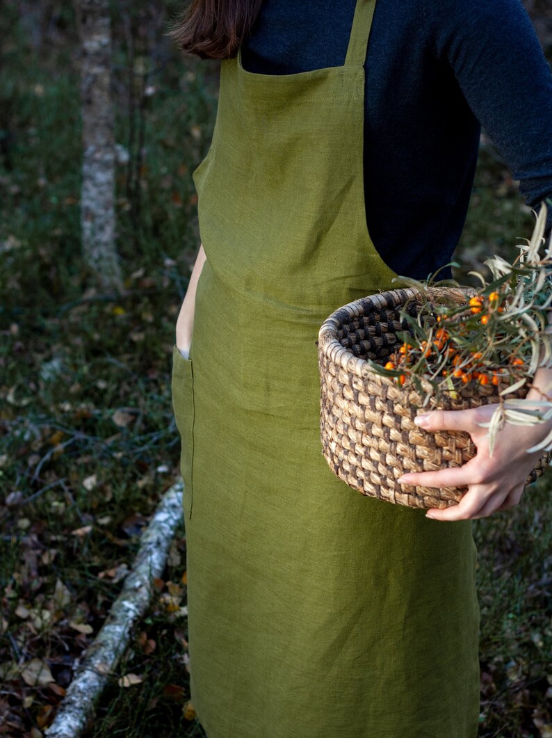 May include: A person wearing a green linen apron with a large pocket on the left side, holding a woven basket filled with orange berries and green leaves.