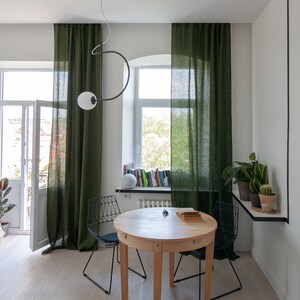 May include: A round wooden dining table with two black metal chairs. The table is set with a book and a pair of glasses. The room has green linen curtains, a window with a view of trees, and a shelf with books.