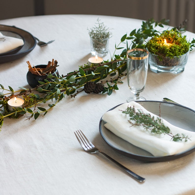 May include: A white tablecloth with a green garland of leaves and branches, a small glass vase with a plant, a small black bowl with cinnamon sticks, a glass of water, a plate with a white napkin and a sprig of greenery, a fork, and two small candles.