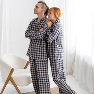 May include: Matching navy and beige plaid pajama sets. Each set includes a long-sleeved button-down shirt and pants. The pajamas are worn by two people, standing near a white chair and a woven rug. The background is a white wall and sheer curtains.
