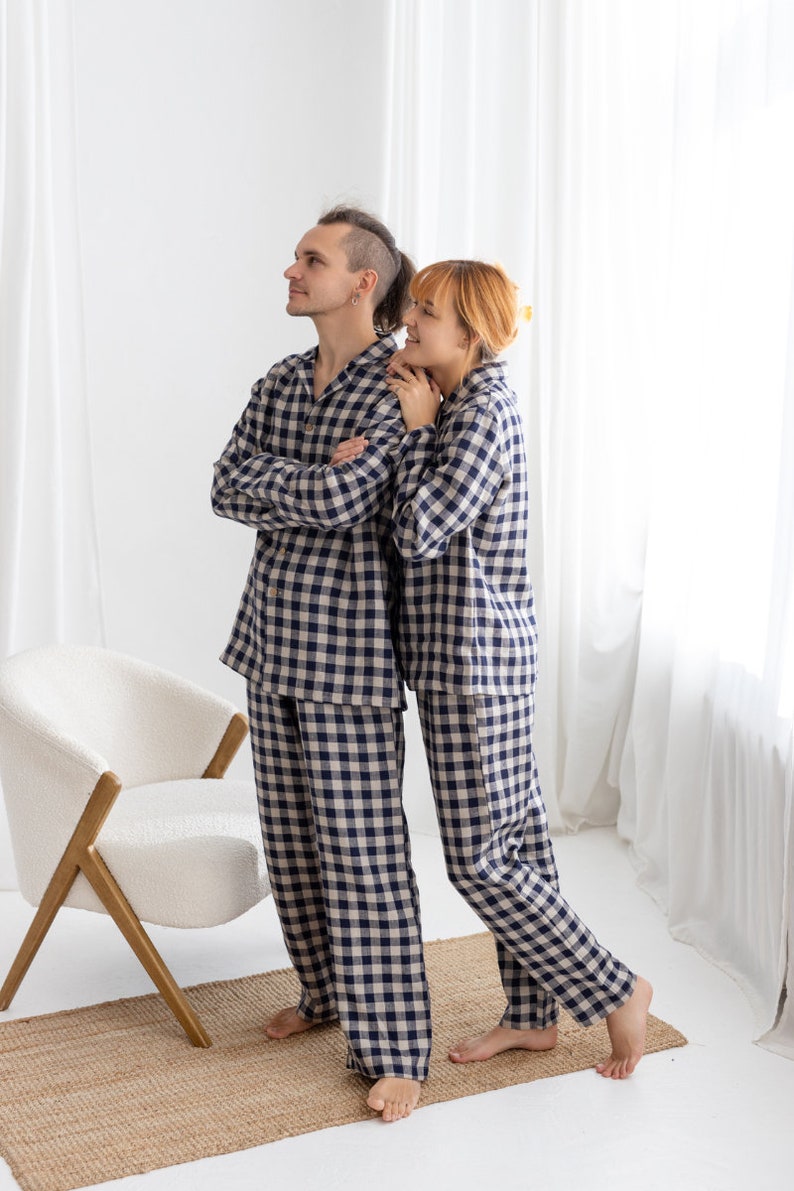 May include: Matching navy and beige plaid pajama sets. Each set includes a long-sleeved button-down shirt and pants. The pajamas are worn by two people, standing near a white chair and a woven rug. The background is a white wall and sheer curtains.