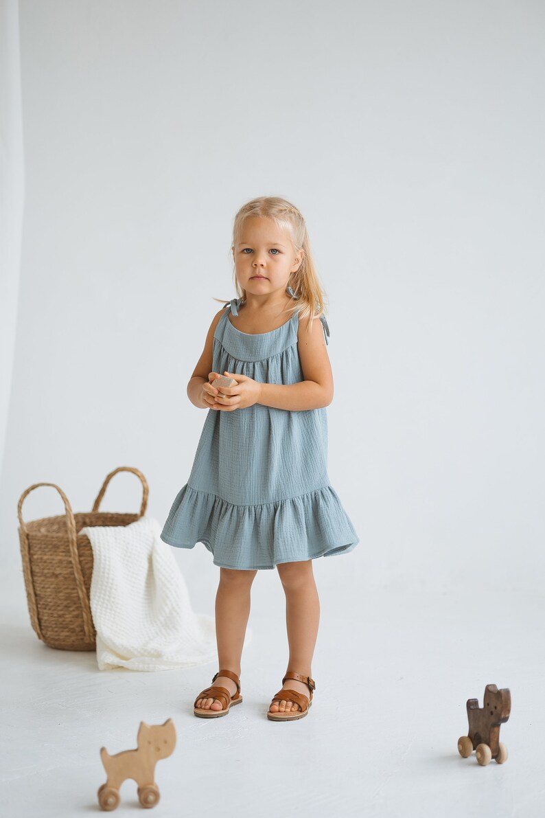 May include: A young child wearing a light blue dress with a ruffled hem and brown leather sandals. The dress has tie straps. A woven basket and wooden toys are in the background.