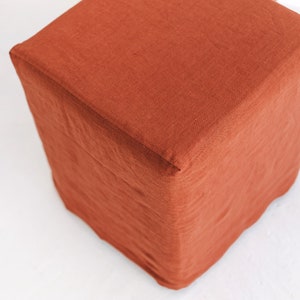 May include: A square, terracotta-coloured linen ottoman with a smooth, textured surface.