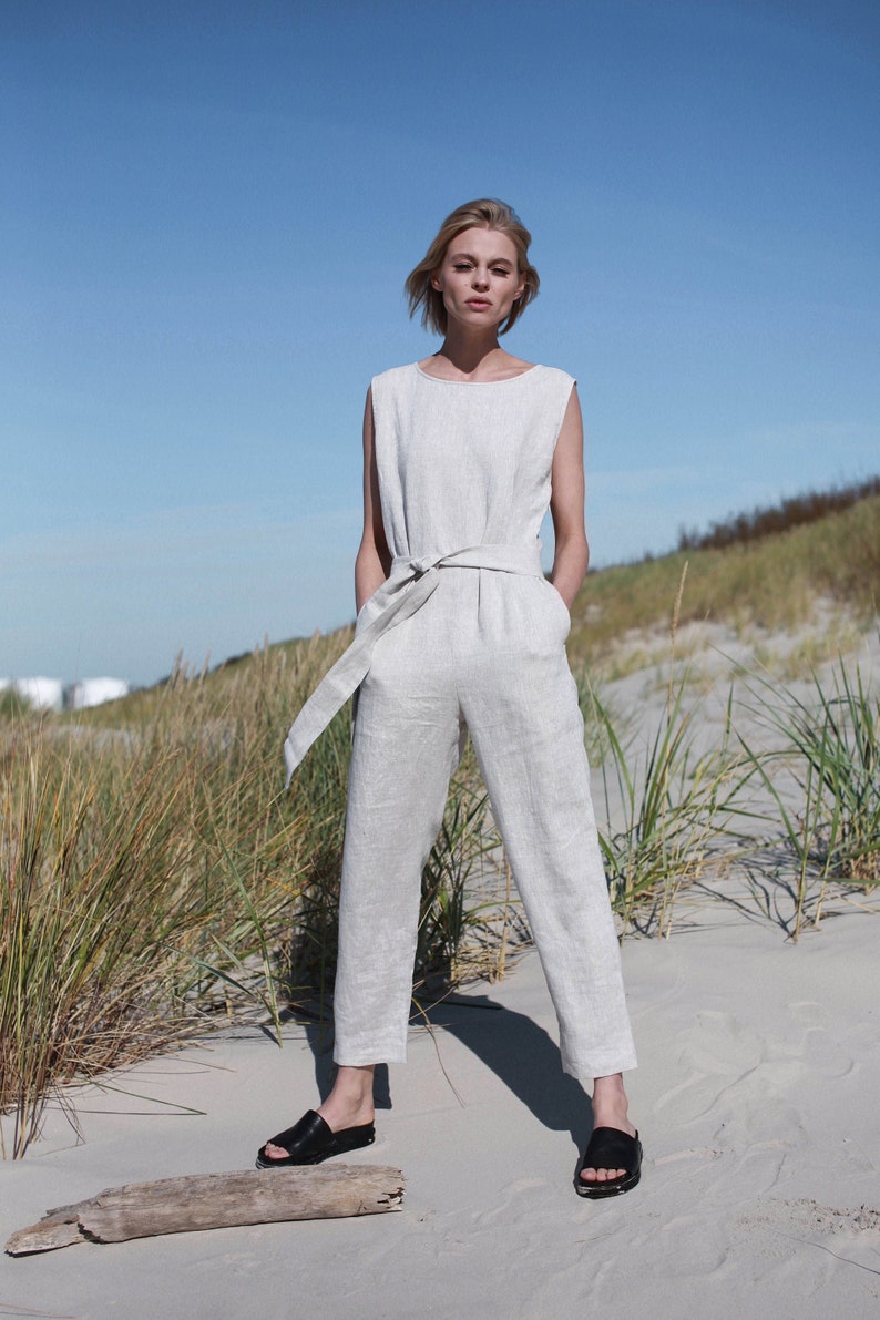 May include: A woman wearing a light gray linen jumpsuit with a wide belt and black sandals. She is standing on a sandy beach with tall grass in the background.