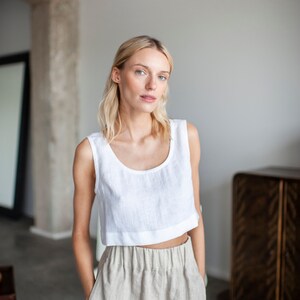 K&ouml;nnte beinhalten: Ein wei&szlig;es Leinen-Crop-Top mit Rundhalsausschnitt und eine hellbeige Leinenhose. Das Outfit ist schlicht und elegant, perfekt f&uuml;r einen l&auml;ssigen, aber stilvollen Look. Das Model tr&auml;gt das Outfit in einem hellen Raum.