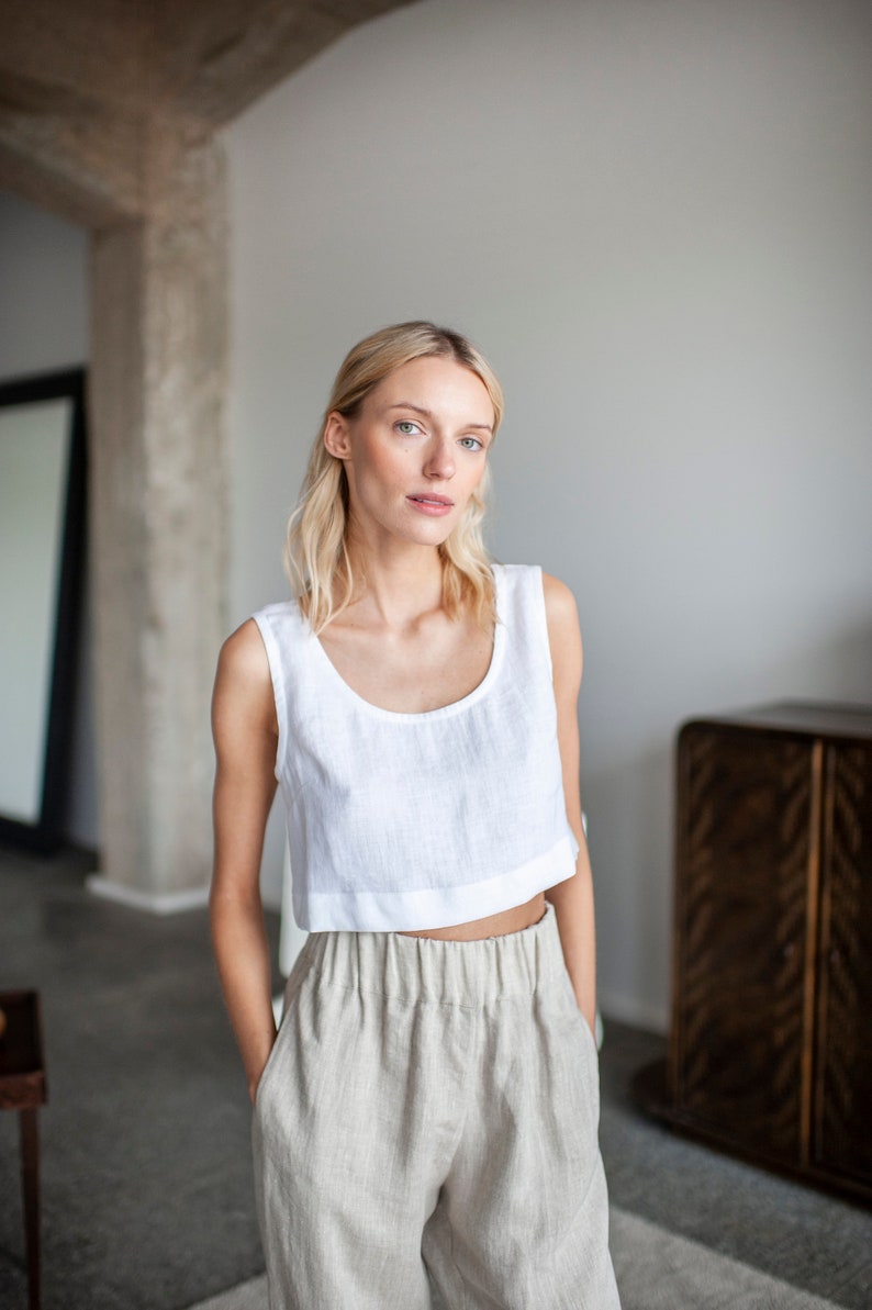K&ouml;nnte beinhalten: Ein wei&szlig;es Leinen-Crop-Top mit Rundhalsausschnitt und eine hellbeige Leinenhose. Das Outfit ist schlicht und elegant, perfekt f&uuml;r einen l&auml;ssigen, aber stilvollen Look. Das Model tr&auml;gt das Outfit in einem hellen Raum.