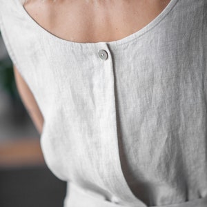 May include: A white linen dress with a single button closure in the back. The dress has a simple, relaxed fit and a tie belt at the waist.