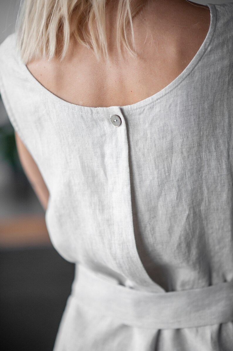 May include: A white linen dress with a single button closure in the back. The dress has a simple, relaxed fit and a tie belt at the waist.