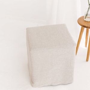 May include: A square, light beige linen ottoman with a textured surface.