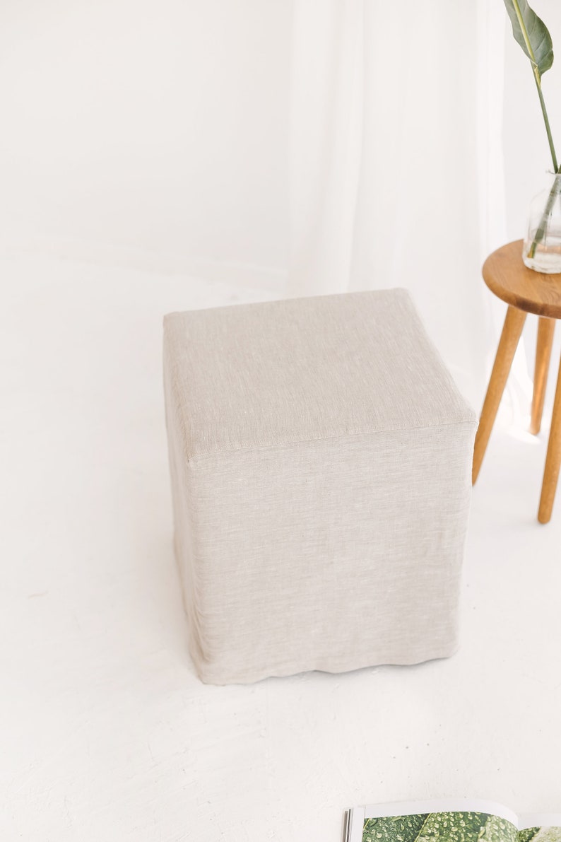 May include: A square, light beige linen ottoman with a textured surface.