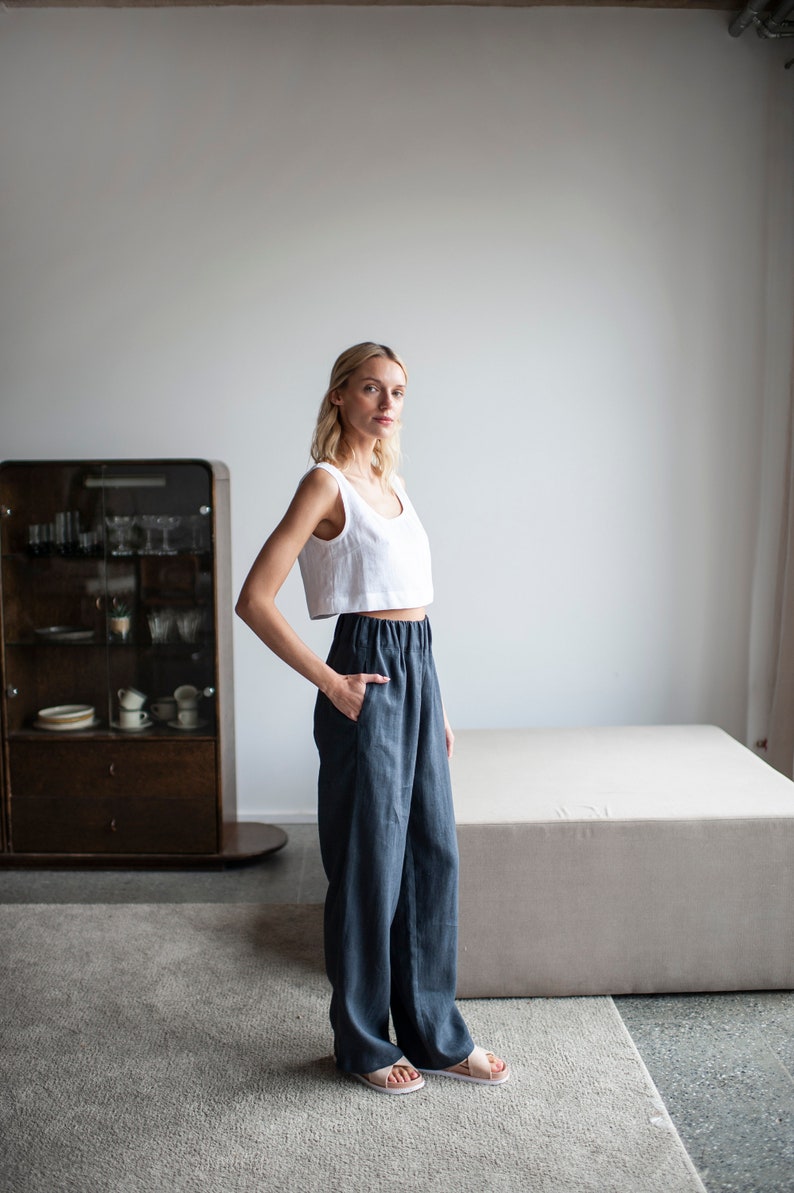 K&ouml;nnte beinhalten: Eine Frau tr&auml;gt ein wei&szlig;es Crop-Top und eine dunkelblaue Leinenhose. Die Hose hat eine lockere Passform mit einem elastischen Bund. Das Outfit wird mit hellfarbenen Sandalen kombiniert. Der Hintergrund umfasst einen Schrank und eine neutralfarbene Plattform.