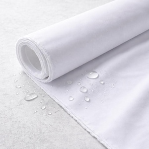 May include: A roll of white fabric with a textured surface, partially unrolled to reveal the inner layers. Water droplets are scattered across the fabric, suggesting a waterproof or water-resistant quality. The fabric's edge is visible.