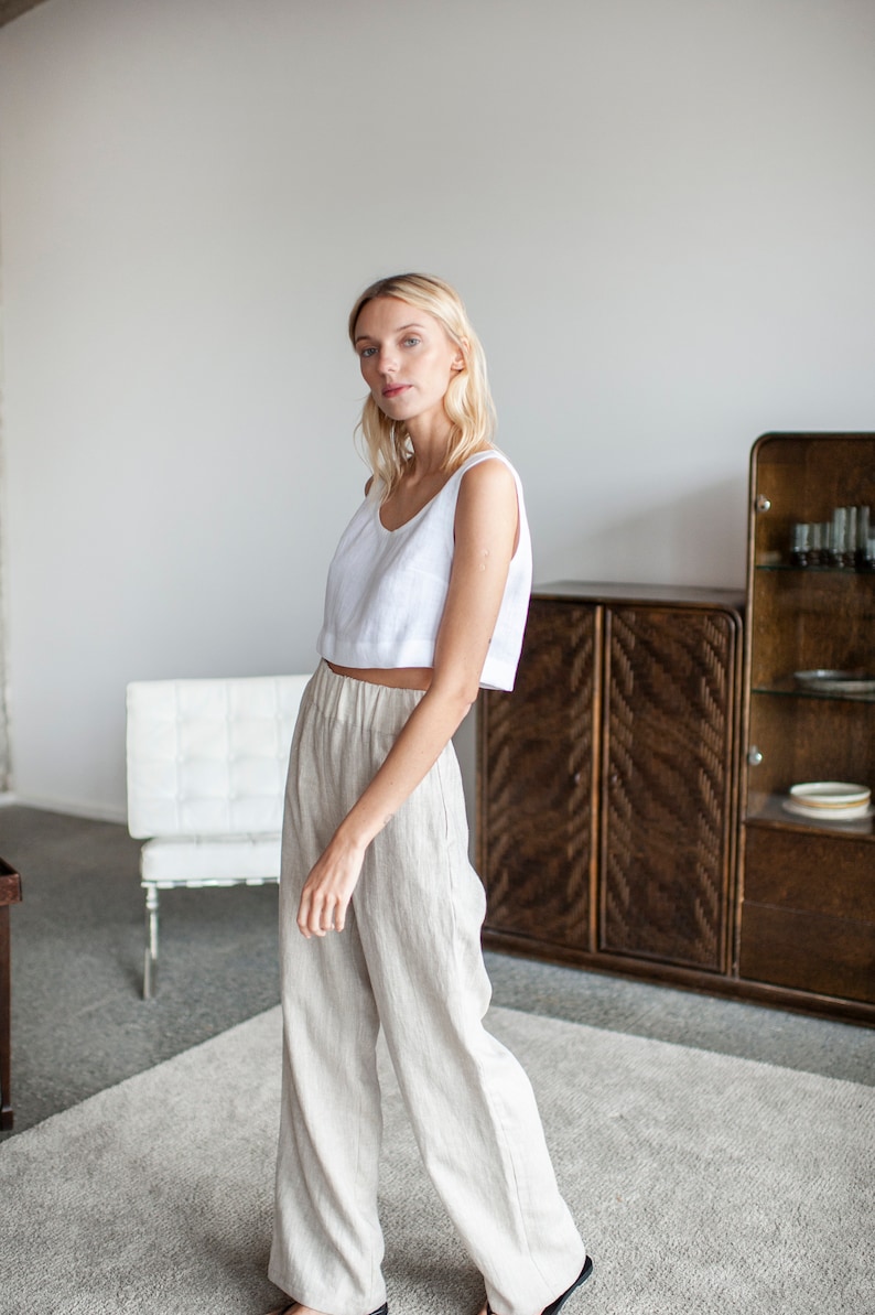 May include: A woman wearing a white linen tank top and beige linen trousers. She is standing in a room with a white wall and a wooden cabinet.