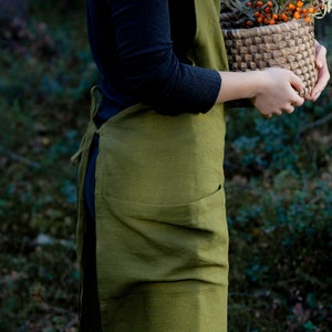 May include: A person wearing a green linen apron with a large pocket. They are holding a woven basket filled with orange berries and green leaves.