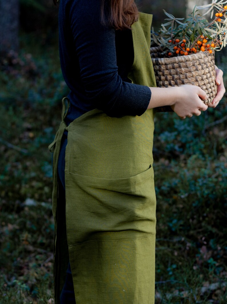 May include: A person wearing a green linen apron with a large pocket. They are holding a woven basket filled with orange berries and green leaves.