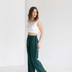 K&ouml;nnte beinhalten: Eine Frau tr&auml;gt ein wei&szlig;es Crop-Top und eine hoch taillierte, weit geschnittene, dunkelgr&uuml;ne Hose. Dazu cremefarbene Sandalen. Der Hintergrund ist eine schlichte wei&szlig;e Wand.
