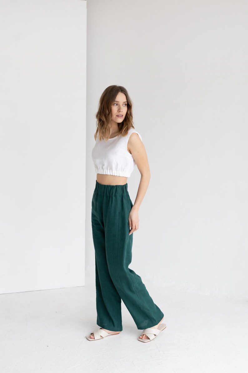 K&ouml;nnte beinhalten: Eine Frau tr&auml;gt ein wei&szlig;es Crop-Top und eine hoch taillierte, weit geschnittene, dunkelgr&uuml;ne Hose. Dazu cremefarbene Sandalen. Der Hintergrund ist eine schlichte wei&szlig;e Wand.