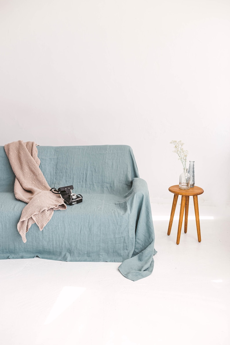 May include: A light blue linen sofa with a beige throw blanket and a vintage camera on the seat. A small wooden side table with a vase of white flowers sits next to the sofa.