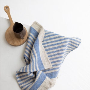 May include: A blue and white striped linen tea towel folded on a white surface. A wooden cutting board with a brown pitcher on top is in the background.