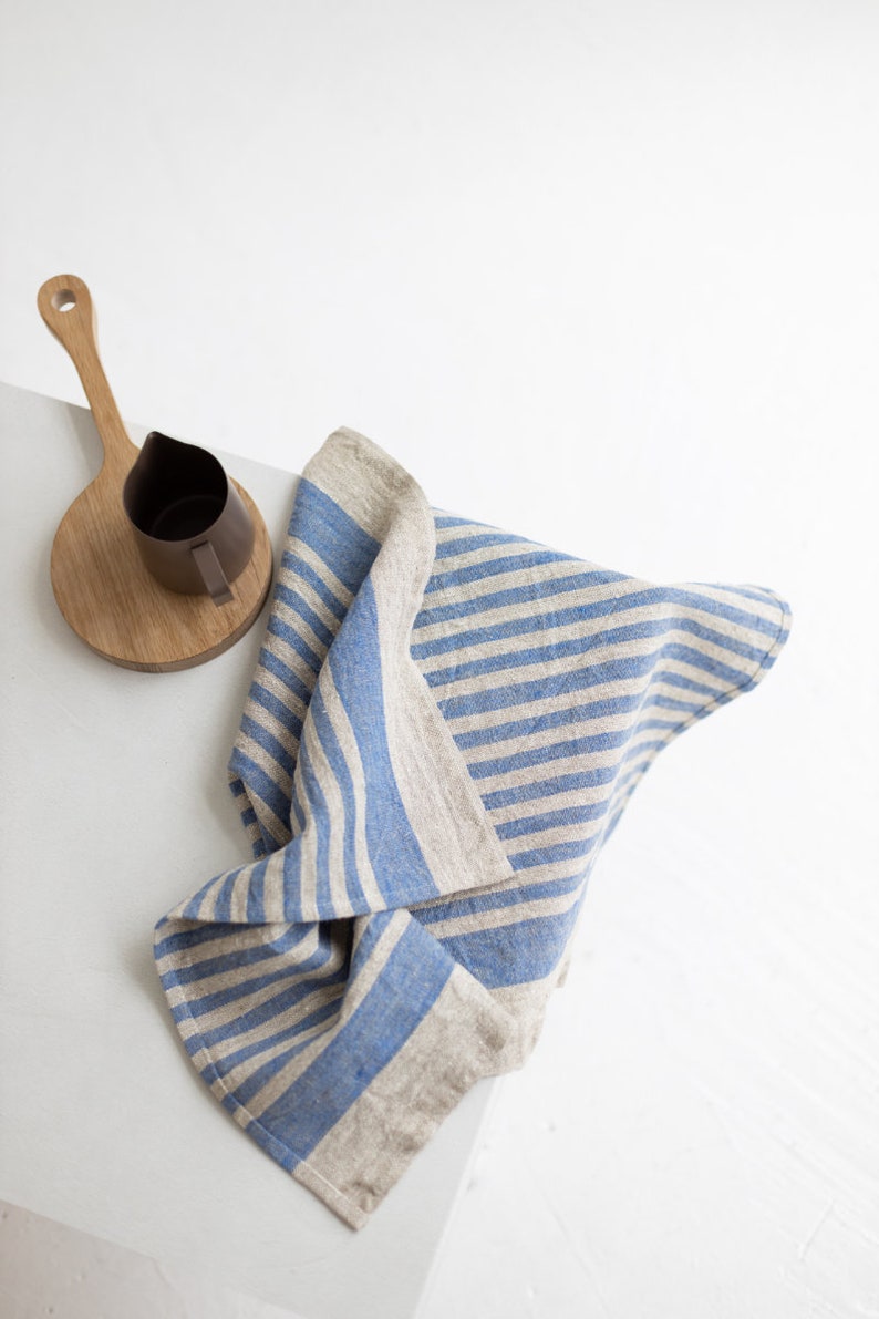 May include: A blue and white striped linen tea towel folded on a white surface. A wooden cutting board with a brown pitcher on top is in the background.