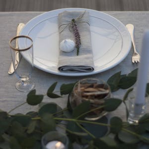 Pu&ograve; includere: Un piatto bianco con un tovagliolo di lino grigio legato con uno spago e un rametto di fiori viola. Un meringa bianca si trova sul tovagliolo. Il piatto &egrave; posto su una tovaglia grigia con un bicchiere d'acqua e una forchetta.