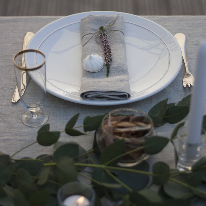 Pu&ograve; includere: Un piatto bianco con un tovagliolo di lino grigio legato con uno spago e un rametto di fiori viola. Un meringa bianca si trova sul tovagliolo. Il piatto &egrave; posto su una tovaglia grigia con un bicchiere d'acqua e una forchetta.