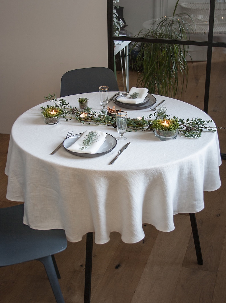 May include: A white linen tablecloth with a round shape, set for a dinner party. The table is decorated with a garland of greenery and small glass candle holders. Two place settings are visible, each with a white napkin, a gray plate, a fork, and a knife.