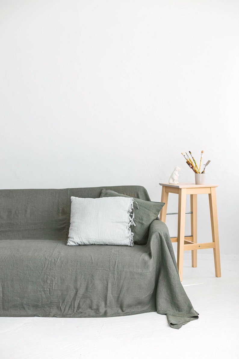 May include: A green linen sofa with a white pillow and a wooden stool with a white ceramic figurine and paintbrushes in a glass jar on top.