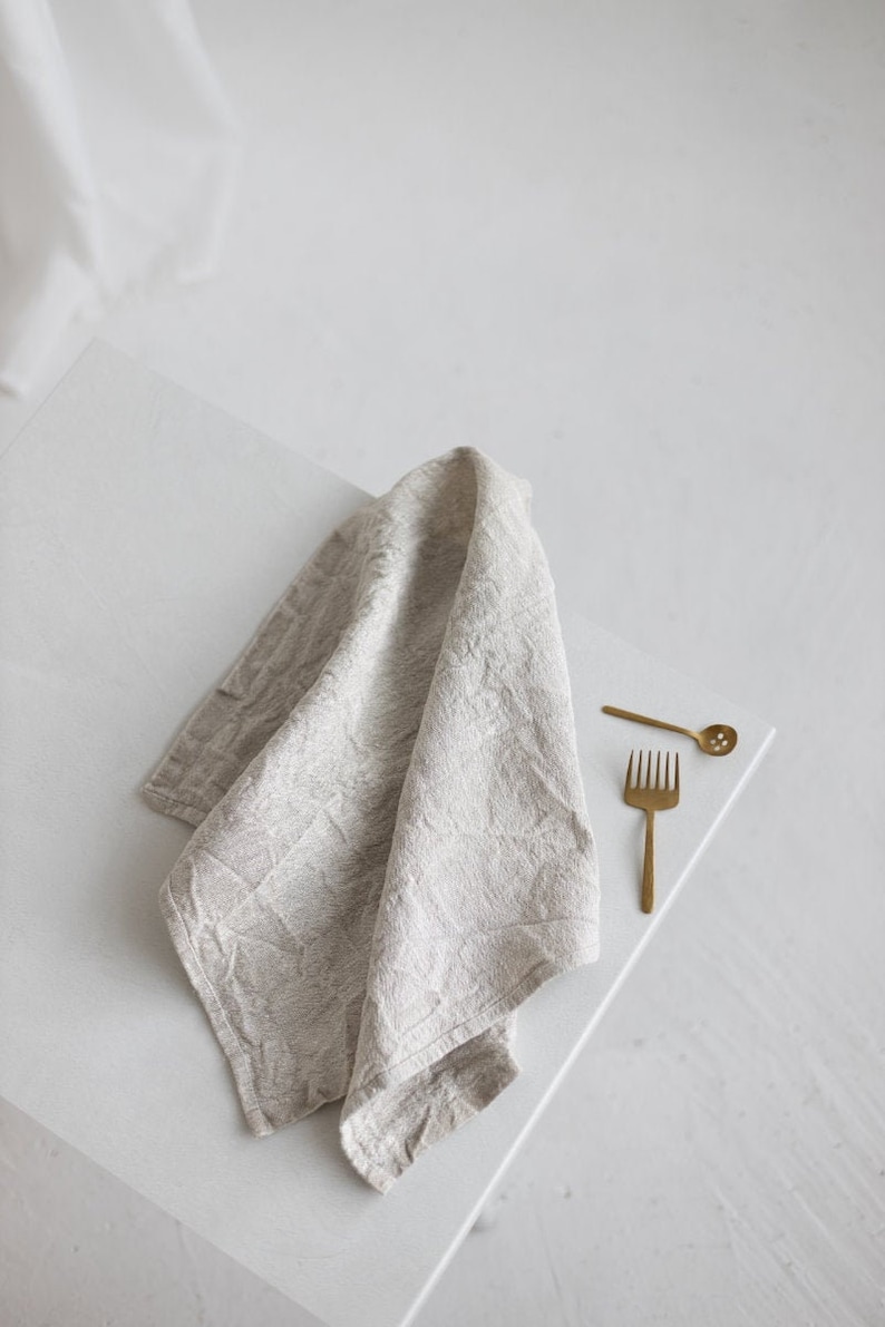 May include: A light gray linen kitchen towel folded on a white surface with two gold metal utensils, a fork and a slotted spoon.