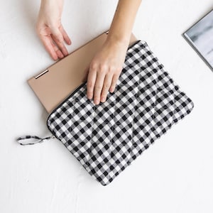 May include: Black and white gingham laptop sleeve with a zipper closure and a handle. The sleeve is quilted and has a soft, padded interior to protect your laptop.