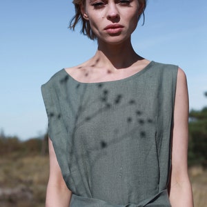 May include: A woman wearing a sage green linen dress with a tie waist. The dress has a relaxed fit and a sleeveless design.