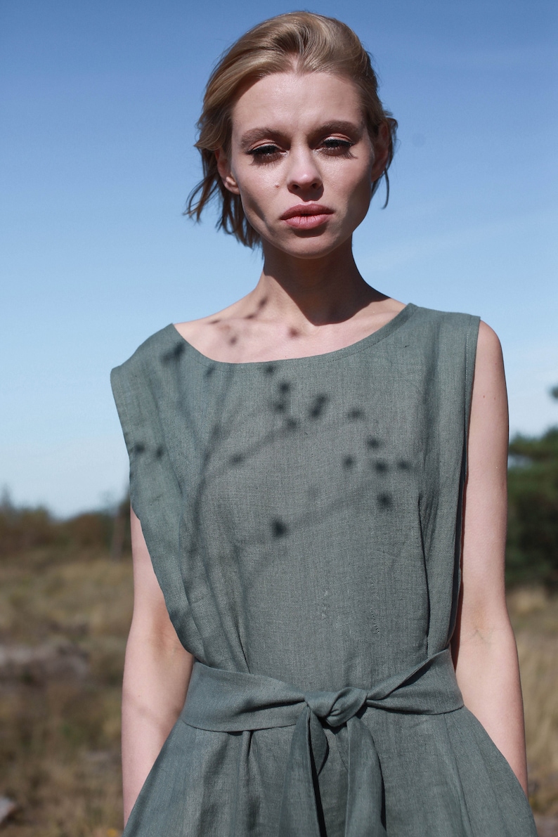 May include: A woman wearing a sage green linen dress with a tie waist. The dress has a relaxed fit and a sleeveless design.