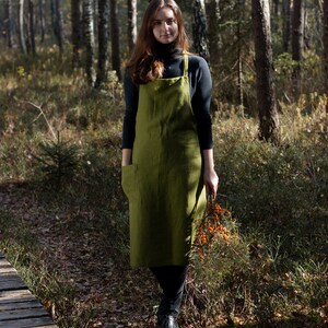 May include: A woman wearing a green linen apron stands in a forest setting. She is holding a branch of orange berries in her right hand.