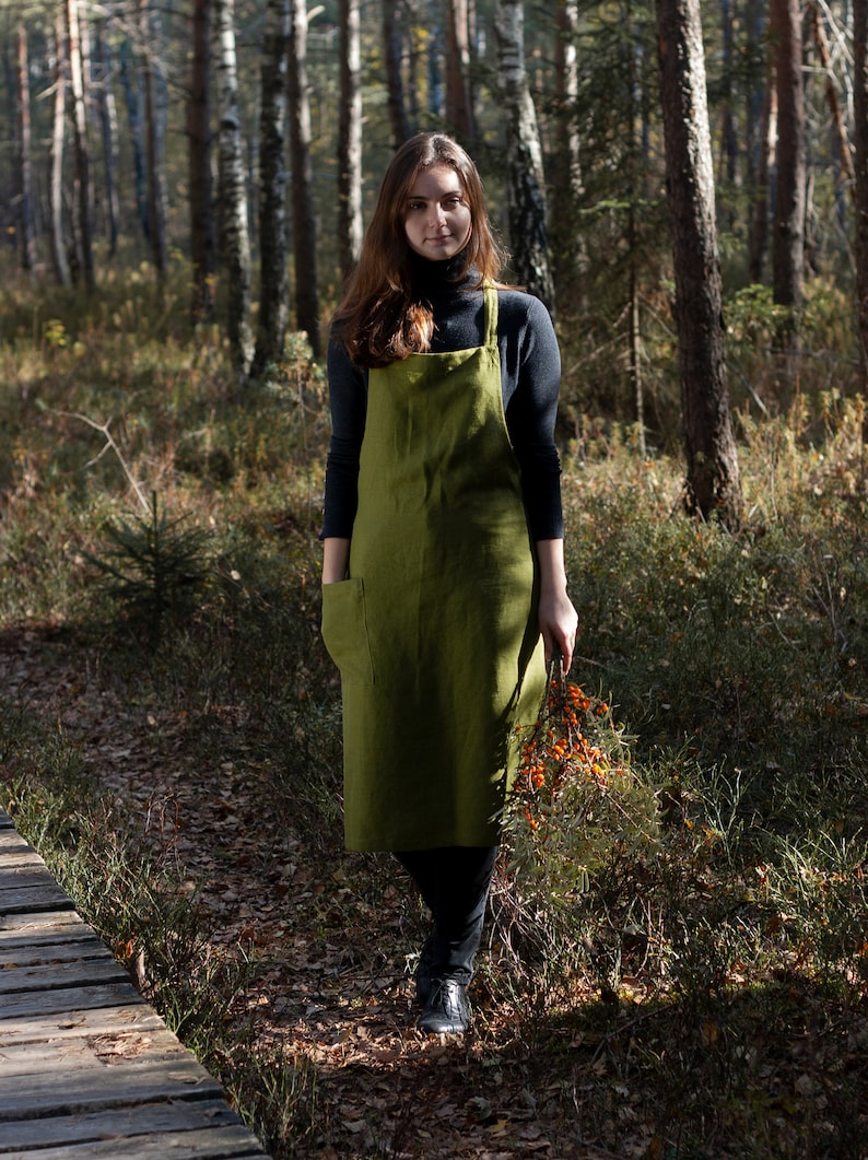 May include: A woman wearing a green linen apron stands in a forest setting. She is holding a branch of orange berries in her right hand.