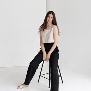 K&ouml;nnte beinhalten: Eine Frau tr&auml;gt ein beiges Crop-Top und eine schwarze Hose und sitzt auf einem schwarzen Hocker. Das Outfit wird mit cremefarbenen Sandalen kombiniert. Der Hintergrund ist eine schlichte wei&szlig;e Wand.