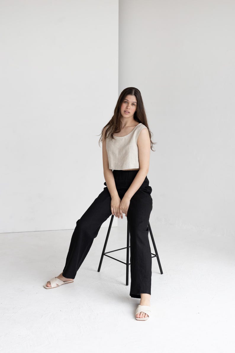 K&ouml;nnte beinhalten: Eine Frau tr&auml;gt ein beiges Crop-Top und eine schwarze Hose und sitzt auf einem schwarzen Hocker. Das Outfit wird mit cremefarbenen Sandalen kombiniert. Der Hintergrund ist eine schlichte wei&szlig;e Wand.